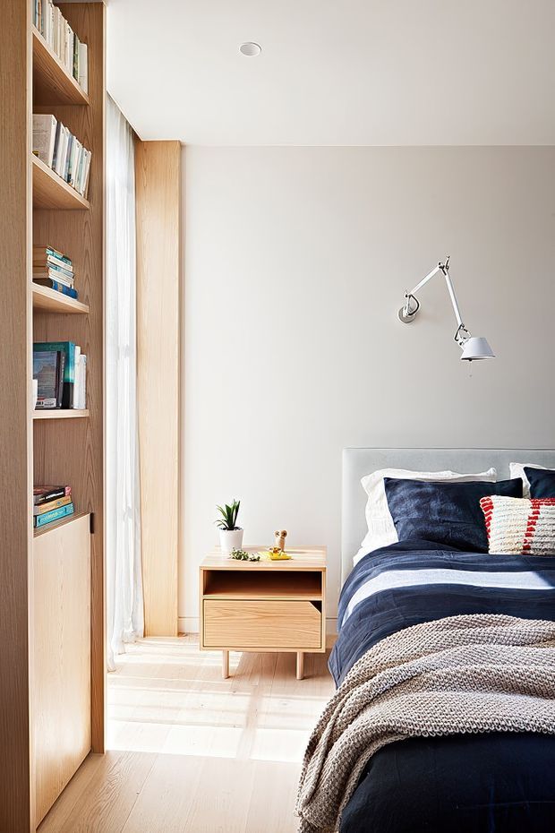 A bright, refined bedroom with a built-in timber bookshelf along the left wall, pale timber flooring and window trim, and soft white walls. The room features a neatly made bed with navy blue bedding, white pillows, a textured neutral throw, and a small red-and-white accent cushion. A compact timber bedside table with drawers sits beside the bed, while a wall-mounted articulated reading lamp and sheer floor-length curtains complete the calm, minimalist interior.