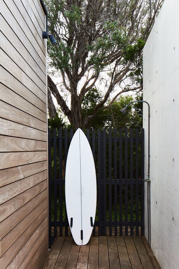 A narrow outdoor passage or surf-access courtyard between a horizontal timber-clad wall and a smooth white rendered wall. A white surfboard leans against a dark vertical-slat gate, with an outdoor shower mounted on the right wall. Timber decking covers the ground, and mature greenery is visible beyond the gate.