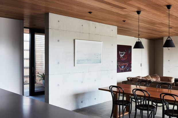 Open-plan contemporary dining area viewed from the kitchen, featuring a long timber dining table with black bentwood-style chairs, a pale exposed concrete feature wall, warm timber-lined ceiling, black pendant lights, dark flooring, and glimpses of an adjoining outdoor area through full-height glazing.