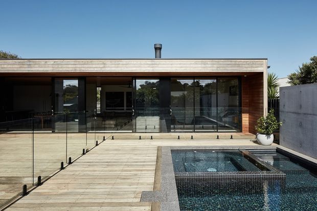 A low, flat-roofed contemporary home opens onto a timber deck and a tiled swimming pool. Full-height dark-framed glazing spans the facade, with warm timber lining around the recessed openings, a concrete boundary wall, glass pool balustrade, and a small chimney visible above the roofline. The image is photographed from the outdoor pool deck, looking toward the house.