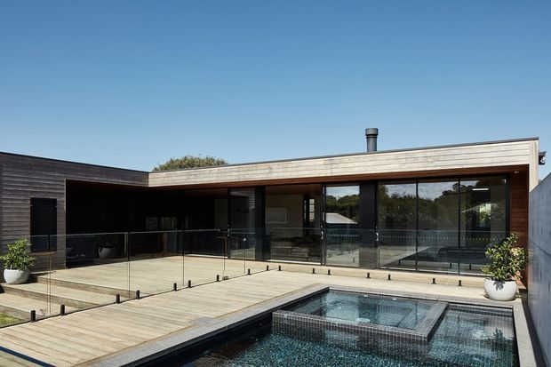 Exterior view of a low-profile contemporary home with a flat roof, dark horizontal cladding, timber-lined soffits and facade elements, extensive floor-to-ceiling glazing, and a timber deck surrounding a tiled swimming pool and raised spa. Glass balustrades frame the deck, with planted pots and a clear blue sky completing the setting.