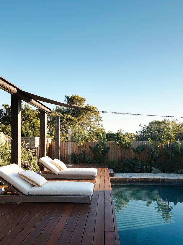 A sunlit outdoor pool area with a dark timber deck, two cushioned lounge chairs, a timber pergola, and a rectangular swimming pool bordered by stone coping. Dense tropical-style planting and a timber privacy fence enclose the space, creating a relaxed resort-like setting.