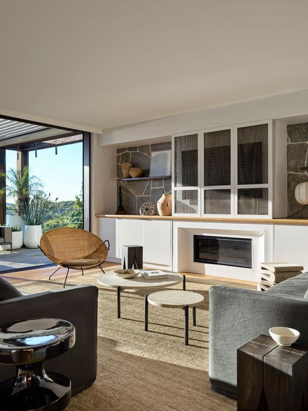Sunlit contemporary living room with a large glazed opening to an outdoor terrace, a stone feature wall, white built-in cabinetry and fireplace, woven lounge chair, neutral sofas, and low round coffee tables. The space has a warm, refined mid-century-inspired feel with strong indoor-outdoor connection.