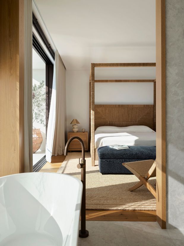 A bright, refined bedroom viewed through an open doorway, with a freestanding white bathtub in the foreground suggesting an adjoining bathroom. The bedroom features a woven timber or rattan four-poster bed with white bedding and a dark navy textured throw, a small bedside table and lamp, floor-to-ceiling glazing with sheer curtains, warm timber flooring, and a natural woven rug. The space has warm sunlight, neutral finishes, and tactile natural materials.