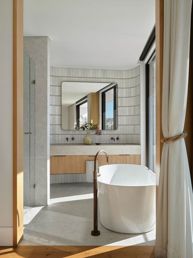 A bright, refined contemporary bathroom featuring a freestanding white bathtub beside full-height windows dressed with cream curtains. The room includes a wall-mounted timber vanity with a pale stone benchtop, vertically lined light tiles with curved horizontal grout detailing, a frameless glass shower enclosure, and a rounded rectangular mirror. A dark bronze freestanding bath tap provides contrast, while warm timber framing and natural daylight soften the space.