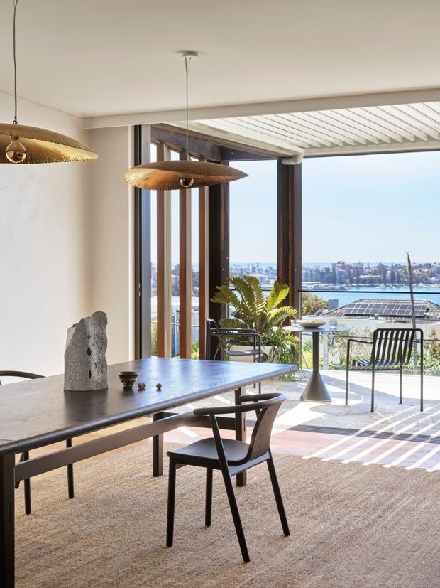 Bright contemporary dining area opening onto a covered terrace, with a long dark dining table, sculptural pendant lights, and black-framed chairs. Large glazed openings provide expansive views across blue water toward a dense urban waterfront, while a slatted pergola casts linear shadows across the terrace and neutral carpeted interior. Timber-framed glazing, white walls, and indoor greenery create a refined, airy setting.