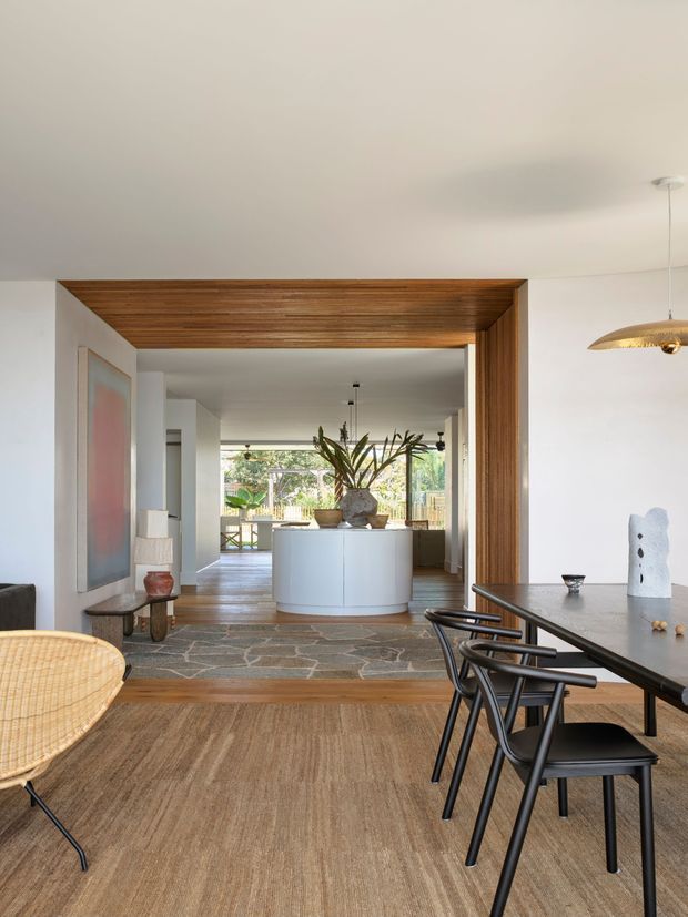 A refined open-plan interior viewed from a dining area toward a bright living and kitchen zone. A broad timber-lined portal frames the opening, with white walls, a natural woven floor covering, irregular stone flooring beyond, a curved white island or reception-like joinery element, indoor plants, and black dining furniture. Large glazing opens the rear of the home to greenery and daylight. The palette is warm, natural, and contemporary with mid-century-inspired furniture and sculptural decor.
