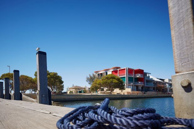 Exterior waterfront view across a calm canal from a timber dock or boardwalk. Thick coiled marine rope and dock posts dominate the foreground, while a contemporary multi-storey residential building with balconies and bold red facade sections is visible across the water under a clear blue sky.