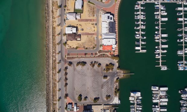 Aerial top-down view of a waterfront marina with rows of moored boats, a large paved parking area, access roads, and adjacent low-rise buildings along the shoreline. The image is dominated by the marina and surrounding waterfront site rather than an individual interior space.