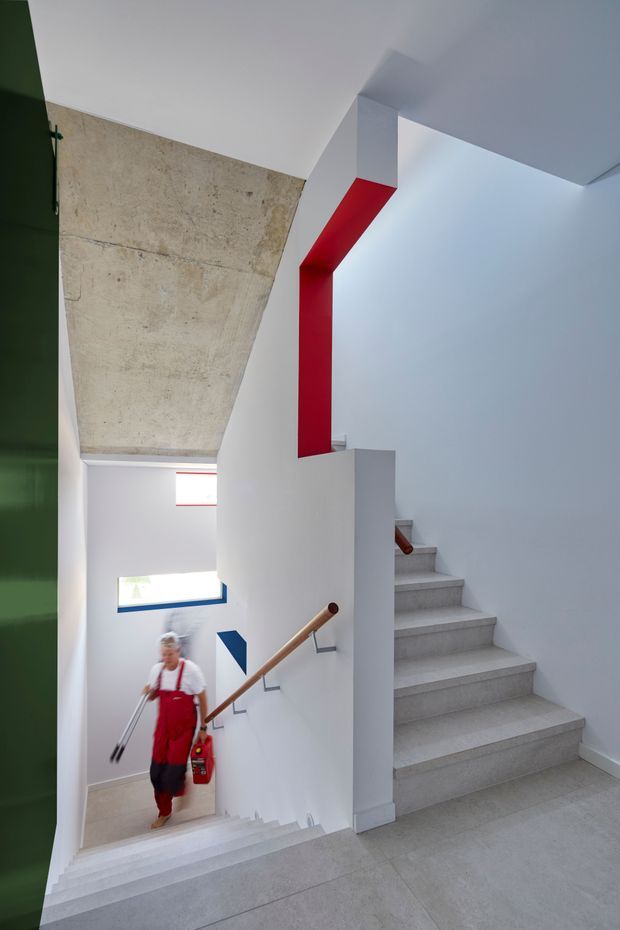 A bright, minimalist stairwell with two flights of pale concrete or stone stairs, white walls, timber handrails, and bold red and blue geometric accents. A large exposed concrete ceiling plane contrasts with the crisp white interior, while a dark green vertical surface frames the left edge. The space has a sculptural, contemporary architectural character and appears to connect multiple levels.