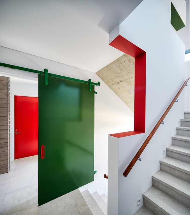 A brightly lit modern stairway and hallway with bold colour accents. Grey concrete stairs rise along the right side beside a white wall with a warm timber handrail. A large dark-green sliding barn door hangs from exposed green hardware, while vivid red door and red-painted architectural framing create strong graphic contrast. Pale concrete or stone flooring and a textured light stone wall surface are visible around the stair opening.