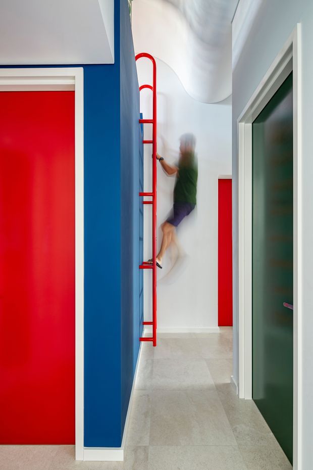 A narrow, boldly coloured interior corridor with vivid blue walls, red doors and a bright red wall-mounted ladder-like climbing feature. White trim, pale tiled flooring and a sculptural curved white ceiling create a playful contemporary setting. A blurred person is climbing the red fixture in the distance, while a dark green door is visible on the right.