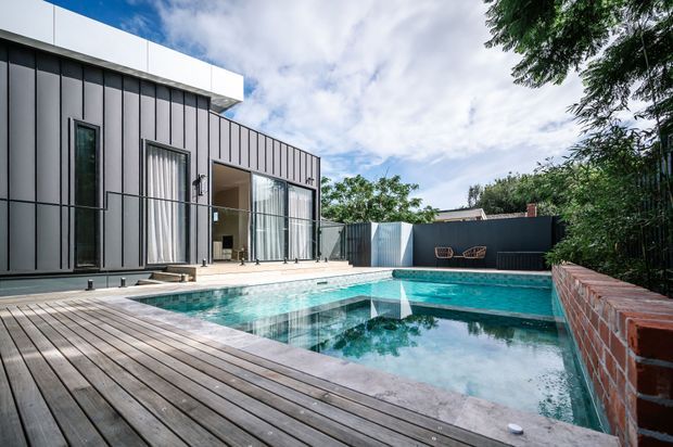 Contemporary residential courtyard viewed from a timber deck, featuring a rectangular swimming pool, dark vertically detailed cladding, expansive glazed doors, and a brick boundary wall surrounded by mature greenery.