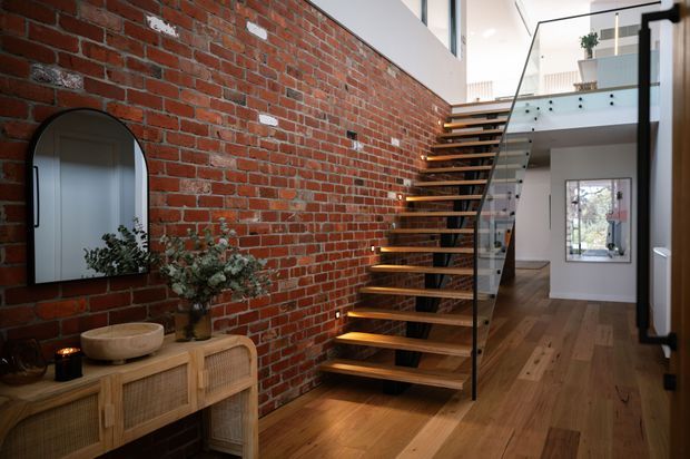 Interior view of a contemporary multi-level entry hall featuring an exposed red brick feature wall, warm timber flooring, and an open-riser timber staircase with a clear glass balustrade and dark metal supports. A rattan console, arched mirror, and potted greenery sit against the brick wall, while a bright upper-level landing and hallway are visible beyond.