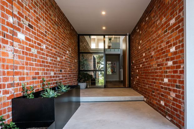 A narrow, covered entry passage with exposed red brick walls, a smooth concrete floor, black metal-framed glazed doors, and a raised threshold leading into the building. Built-in black planter boxes with succulents line one side, while a glimpse of an interior stair or mezzanine is visible beyond the entrance.