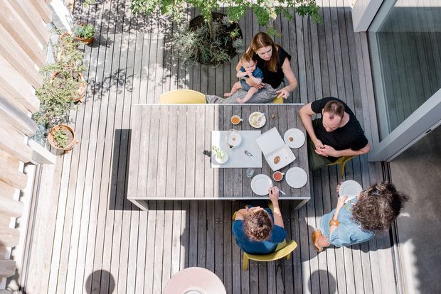 Overhead view of a family gathered around a rectangular outdoor dining table on a timber deck. The deck is bordered by vertical timber screening and potted plants, with large glazed doors opening onto the space. Yellow chairs and tableware add small colour accents to the otherwise natural timber setting.