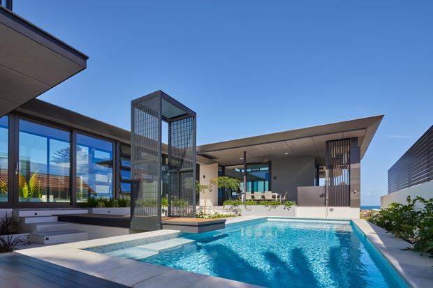 Contemporary flat-roof residence arranged around a large rectangular swimming pool and terrace. The image shows extensive floor-to-ceiling glazing, a tall dark metal screened architectural volume, broad cantilevered roof planes, pale rendered walls, and an open covered outdoor living area with a dining setting. Bright blue sky and low planting frame the modern indoor-outdoor composition.