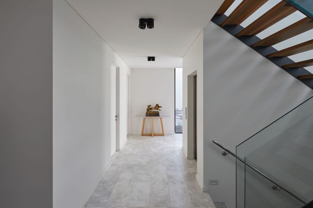 A bright, minimalist residential hallway with white walls and ceiling, pale stone-look tiled flooring, and simple recessed or flush black ceiling lights. A narrow console table with decorative objects sits at the end of the corridor near a glazed opening. To the right, a contemporary staircase features timber treads or beams, a glass balustrade, and a slim metal handrail.