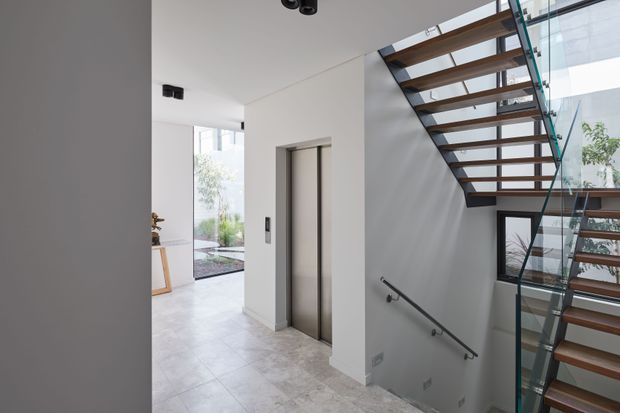 Bright contemporary interior hallway and stair landing with a minimalist open-riser staircase featuring dark timber treads, a slim dark structure, and clear glass balustrades. A stainless-steel elevator door is set into the white wall, while large glazed openings bring in natural light and reveal greenery outside. The floor is finished in pale stone-look tile, and the space has clean lines with understated recessed lighting.