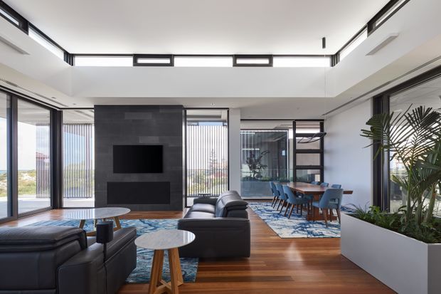A bright, open-plan contemporary living and dining area with a double-height ceiling and extensive floor-to-ceiling glazing. The room features dark leather sofas, round timber side tables, a dark tiled media wall with built-in television and cabinetry, timber flooring, and a dining table with blue upholstered chairs. A large integrated planter separates the living space from the dining area, while slim dark window frames, clerestory glazing, and vertical blinds reinforce the clean architectural character.