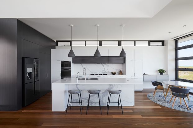 A contemporary open-plan kitchen and dining area with a large white marble-look island, dark full-height cabinetry, integrated appliances, pendant lights, timber flooring, and a small dining setting beside expansive windows. The space is bright and refined, with a view toward a coastal or open landscape beyond the glazing.