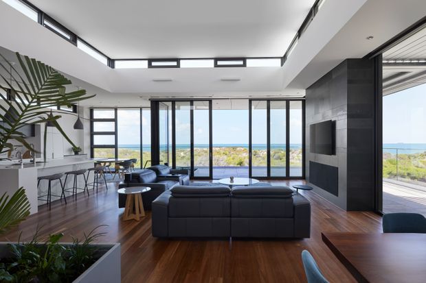Bright open-plan coastal living area with a large black sectional sofa facing a dark tiled fireplace and wall-mounted television. Floor-to-ceiling black-framed glazing opens the room to expansive ocean views, while a kitchen and dining area sit to the left. Warm timber flooring, white walls, indoor plants, and a high clerestory-lit ceiling create an airy contemporary interior.