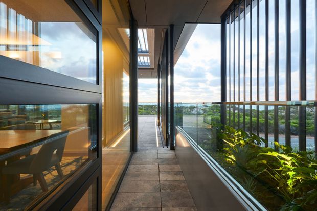 A bright, narrow architectural corridor or covered balcony with floor-to-ceiling glazing, dark metal framing, warm timber-lined walls, and large grey tiled flooring. The space opens toward a glazed balustrade overlooking leafy surroundings and water, with a dining area visible through the glazing on the left. Vertical metal fins and integrated planting create a strong contemporary indoor-outdoor connection.