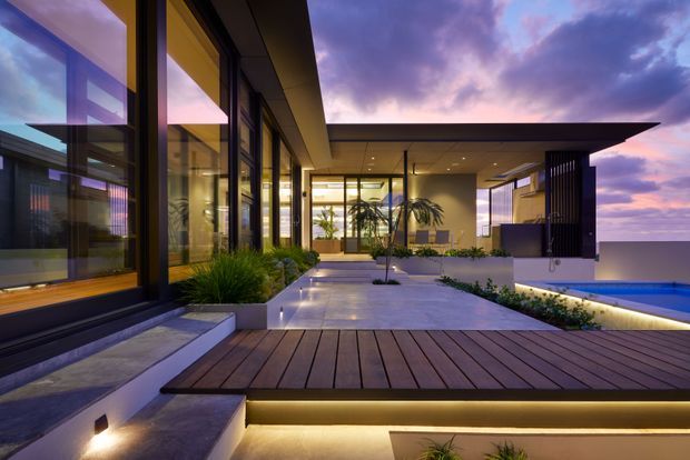 A contemporary single-level residence at dusk, viewed from an outdoor timber deck and paved courtyard. The composition features extensive floor-to-ceiling glazing, dark slender window frames, strong horizontal roof planes, integrated step and deck lighting, landscaped planting beds, and a swimming pool extending to the right. Warm interior light contrasts with the purple-blue evening sky and distant horizon.