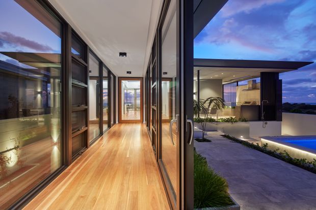 A contemporary residential corridor with warm timber flooring, extensive dark-framed floor-to-ceiling glazing, and built-in timber joinery. The corridor opens directly onto a covered outdoor entertaining area and illuminated swimming pool, with a blue evening sky visible beyond. The image combines a refined interior circulation space with a strong indoor-outdoor connection.