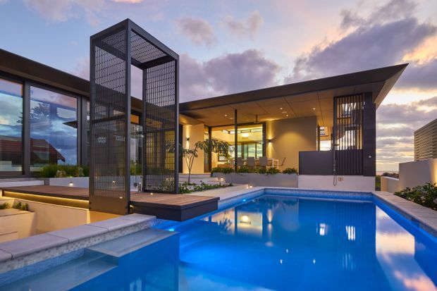 A contemporary single-level residence photographed at dusk, centred on a bright rectangular swimming pool with an integrated shallow ledge and raised spa-like seating area. The home has broad glazing, a deep covered outdoor living area, warm recessed lighting, a projecting flat roof, and tall black metal screens forming a sculptural architectural frame beside the pool. Low planted beds and pale paving surround the pool, with a dramatic cloudy evening sky beyond.