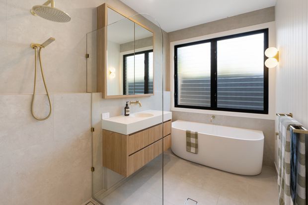 A bright contemporary bathroom featuring a walk-in shower with a frameless glass screen, a floating timber vanity with integrated white basin, a mirrored wall cabinet, and a freestanding white bathtub beneath a large black-framed window. Neutral large-format wall and floor tiles, brushed brass shower and tapware, and a heated towel rail complete the refined design.