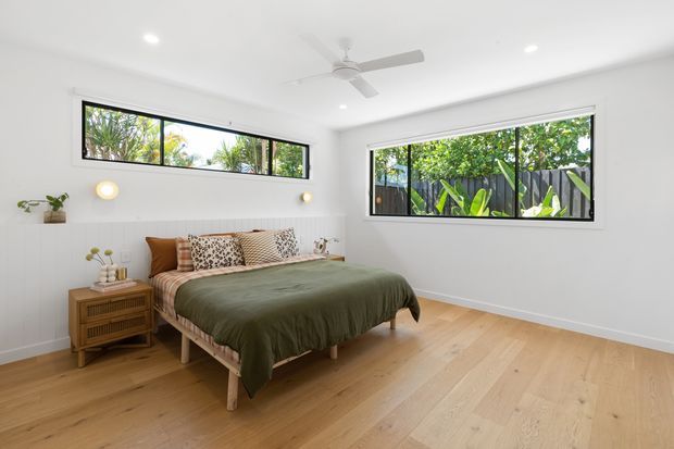 Bright, airy bedroom with white walls and ceiling, a ceiling fan, wide black-framed windows overlooking lush greenery, and pale timber flooring. A low timber bed with muted olive-green bedding, patterned cushions, a woven throw, and a matching timber bedside cabinet creates a relaxed natural feel. Subtle wall sconces and small plants add warmth.