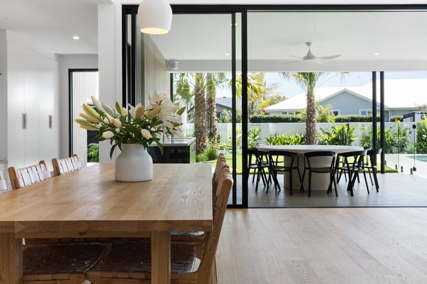 Bright contemporary open-plan dining area with a large light timber dining table in the foreground, woven timber dining chairs, and a white pendant light. Black-framed glazed sliding doors connect the dining space to an outdoor dining area, with a landscaped garden and palm trees visible beyond. White built-in cabinetry and pale timber flooring create a clean, airy interior.