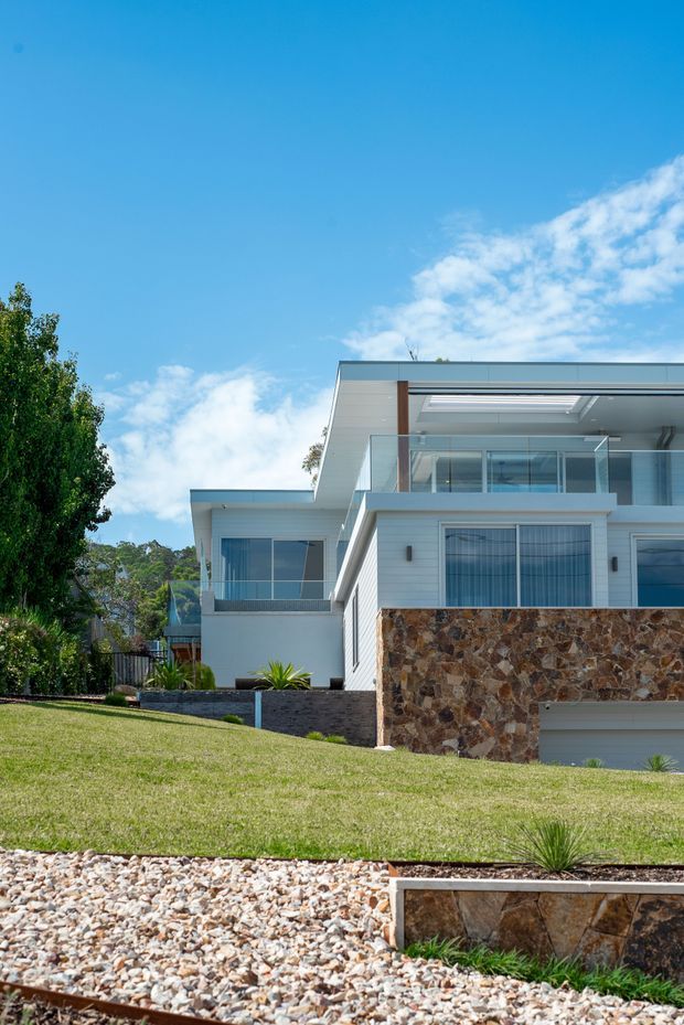 Exterior view of a contemporary multi-level residence on a landscaped sloping site. The home features crisp white horizontal cladding, extensive glazing, glass balcony balustrades, a flat roof with a large roof opening or skylight, and a prominent irregular natural-stone lower facade and retaining wall. A broad lawn, gravel landscaping, and architectural planting surround the house under a bright blue sky.
