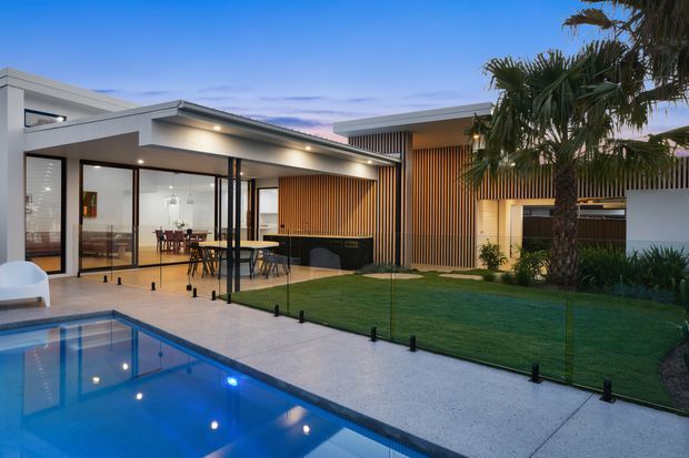 Contemporary single-storey residence viewed from the pool courtyard at dusk. The home features broad floor-to-ceiling glazing, a low-pitched roofline, white rendered walls, warm timber vertical screening, and an open-plan dining area facing the pool. A manicured lawn, palms, frameless glass pool fencing, and pale concrete paving create an indoor-outdoor connection.