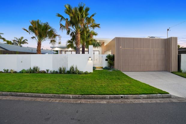 Street-facing view of a contemporary single-storey residence with a flat roof, crisp white boundary walls, a tall vertical timber-look slatted garage door and matching screen, a broad concrete driveway, manicured lawn, and palms in the front garden.