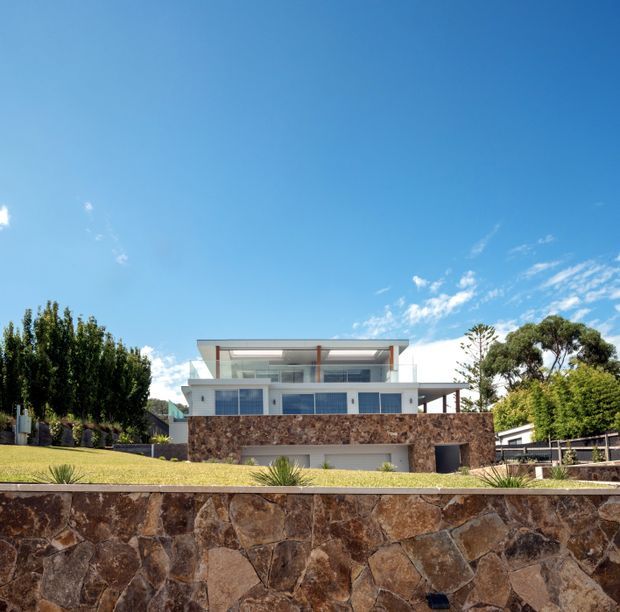 Exterior photograph of a contemporary multi-level residence on a landscaped sloping site. The house has a broad rectilinear form, white rendered walls, extensive glazing, a flat roof, glass balustrades, and dark timber or metal structural accents. Layered random-stone retaining walls dominate the foreground and midground, with manicured planting and mature trees around the property under a clear blue sky.