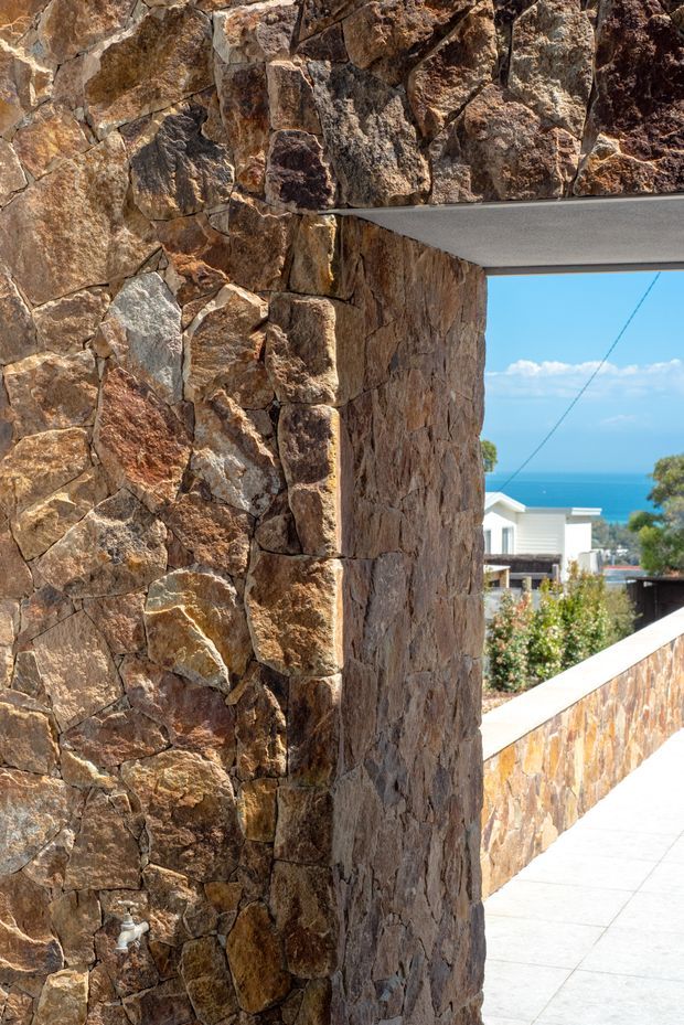 A close architectural detail of a rough, irregular natural-stone wall forming a corner and opening beneath a smooth projecting concrete slab. A low stone-clad boundary wall and pale paved terrace lead toward a bright blue ocean view, with neighbouring coastal buildings visible beyond. The image emphasises textured masonry, the heavy wall junction, and the indoor-outdoor coastal setting.