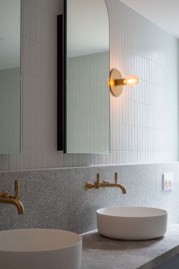 A contemporary bathroom vanity with two round white vessel basins on a grey stone-look countertop. The wall combines vertically grooved white tiles with a grey textured backsplash, complemented by brushed brass wall-mounted taps, warm globe sconces, and large frameless mirrors.