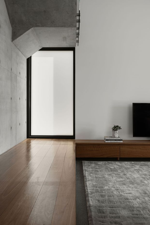 Minimalist living room with a warm timber floor, exposed raw concrete wall and ceiling, black-framed floor-to-ceiling glazing, and a low walnut-toned media console with television. A grey textured rug and small plant add restrained softness to the otherwise monochromatic interior.