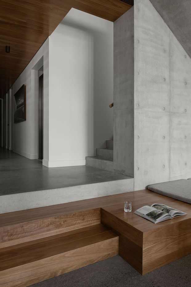 A refined contemporary interior focused on a sunken living area and connecting hallway/stairway. The space features warm timber-clad ceilings and stepped built-in seating or platforms, a large exposed concrete wall and stair structure, smooth white plaster walls, dark grey flooring, and integrated minimalist detailing. A low timber coffee table with an open book sits in the foreground, while a corridor and stairway recede into the background.