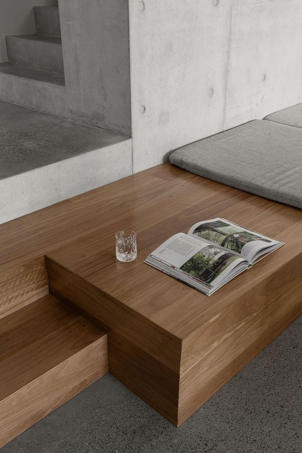 A minimalist concrete interior detail featuring broad concrete steps and walls, a low built-in timber platform or coffee table integrated with stepped seating, grey upholstered cushions, and a small glass with an open magazine. The space has a restrained, warm-neutral palette and strong geometric forms.