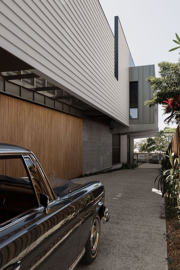 A contemporary multi-level residential exterior viewed from a driveway or narrow access court. The building features crisp projecting volumes, pale horizontal weatherboard-style cladding, dark metal-framed glazing, a tall grey metal-clad upper volume, exposed grey masonry, and a warm vertical timber screen. A vintage car occupies the foreground, while planting and a gated driveway frame the scene.