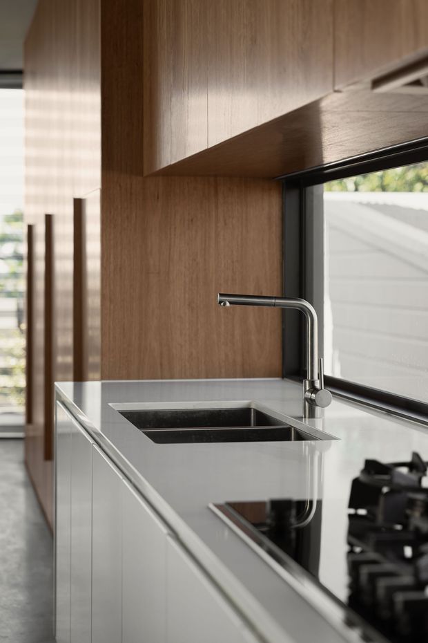 Close-up view of a contemporary kitchen run with a polished light benchtop, integrated black double-bowl sink, brushed metal gooseneck tap, glossy dark cooktop, timber cabinetry and warm timber wall panelling. Black-framed glazing runs alongside the bench, bringing in natural light and views of an adjacent exterior structure.