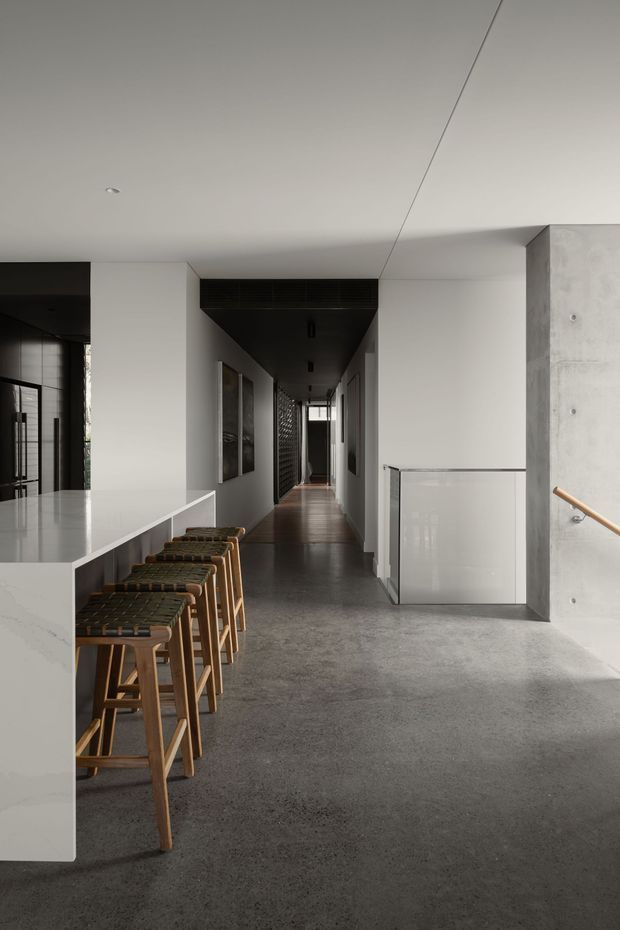 A contemporary open-plan interior viewed from a kitchen or living area toward a long hallway. A white stone-topped island with timber bar stools occupies the left foreground, while the corridor features dark ceiling and wall elements, framed artwork, timber flooring, and glazing at the far end. The right side has a polished concrete wall and a stair opening with a clear glass balustrade and timber handrail. The restrained palette and clean lines create a refined, minimalist atmosphere.