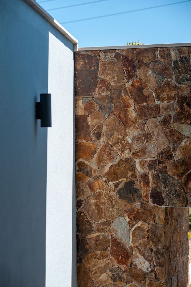 Close-up exterior architectural detail showing a dark grey rendered wall with a minimalist black wall light beside a rough, irregularly jointed natural stone-clad wall. A narrow strip of blue sky and roof edge is visible above, with strong sunlight creating crisp shadows.