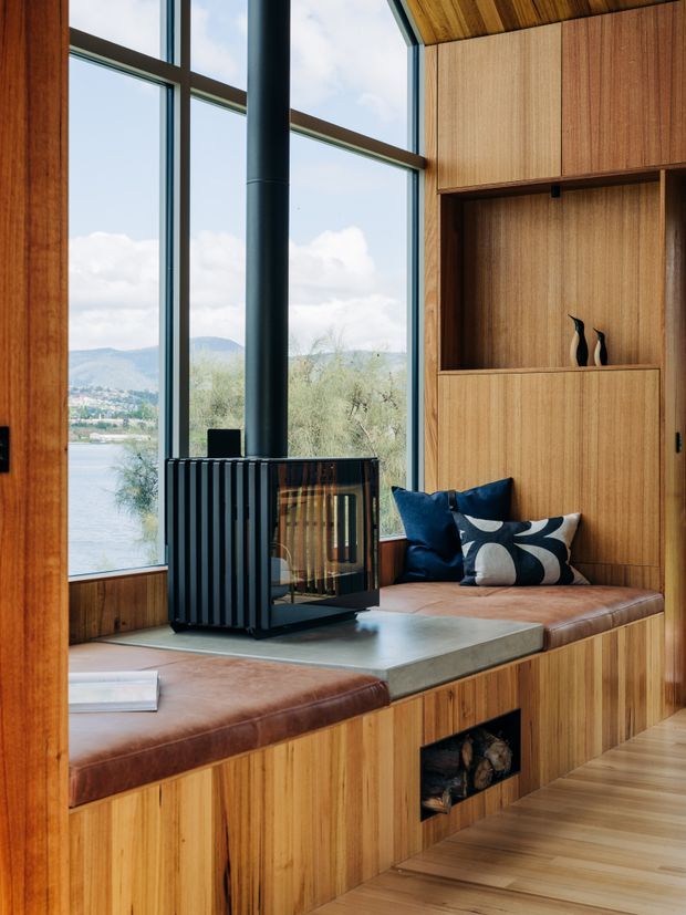 A warm timber-lined interior with a built-in window seat overlooking a lake and hills. A black freestanding wood-burning stove sits on a concrete hearth integrated into the seating platform. Floor-to-ceiling glazing, vertical timber wall panelling, timber cabinetry, leather seat cushions, and dark blue patterned pillows create a cosy contemporary retreat.