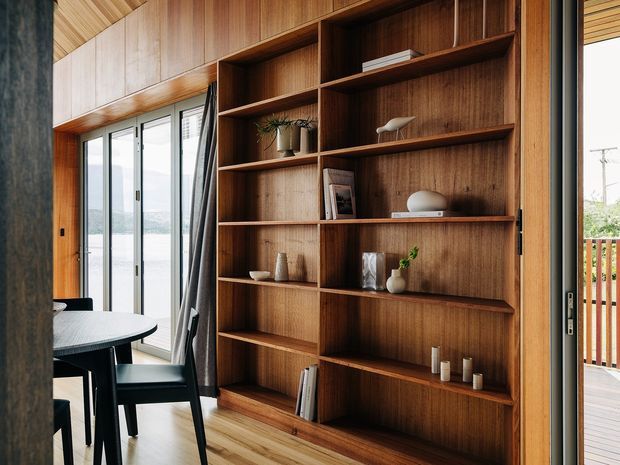 A warm, wood-lined dining and living area with extensive built-in timber shelving and cabinetry. The shelving displays books, ceramics, plants, and decorative objects. A round dining table and black chairs sit beside full-height glazed sliding doors, which open toward a timber deck and exterior view. The ceiling and floor also feature timber finishes, creating a cohesive, natural interior.