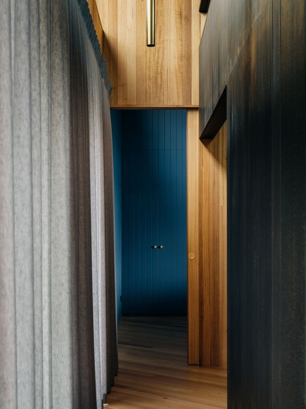A narrow, moody interior passage framed by dark textured wall surfaces and warm vertical timber panelling. A deep blue vertically panelled door or wall is visible at the end, with dark timber flooring, black linear hardware or shelving, and a slim cylindrical pendant light. The composition emphasises layered thresholds, rich timber grain, and strong contrast between blue, natural wood, charcoal, and grey.