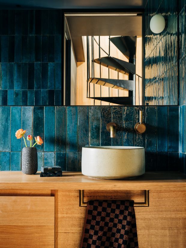 A richly detailed bathroom vanity with deep blue textured wall tiles, a warm timber floating cabinet, a round cream vessel basin, and brass wall-mounted tapware. A mirror reflects a sculptural spiral stairway and timber-clad interior beyond. Strong natural light creates a warm, dramatic atmosphere, with a small vase of flowers and folded textile adding styling.